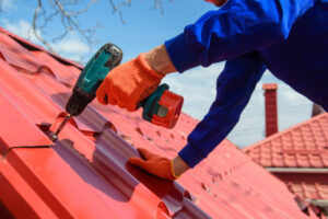 Metal roof installation in Brooklyn NY: Worker securing red metal roofing sheets with a power drill for a durable, energy-efficient roof
