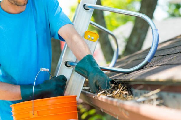 A man in a blue shirt and gloves cleans a roof gutter, ensuring proper drainage and maintenance.
