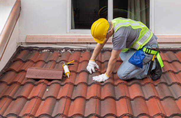 A man in a hard hat and yellow vest replaces shingles on a roof, demonstrating safety and focus on his work.