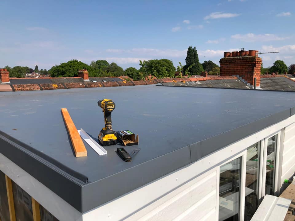 A grey GRP (fiberglass) flat roof under construction, with a yellow power drill, wooden batten, and tools resting on the surface. The background shows a neighborhood of brick houses with traditional chimneys under a clear blue sky.