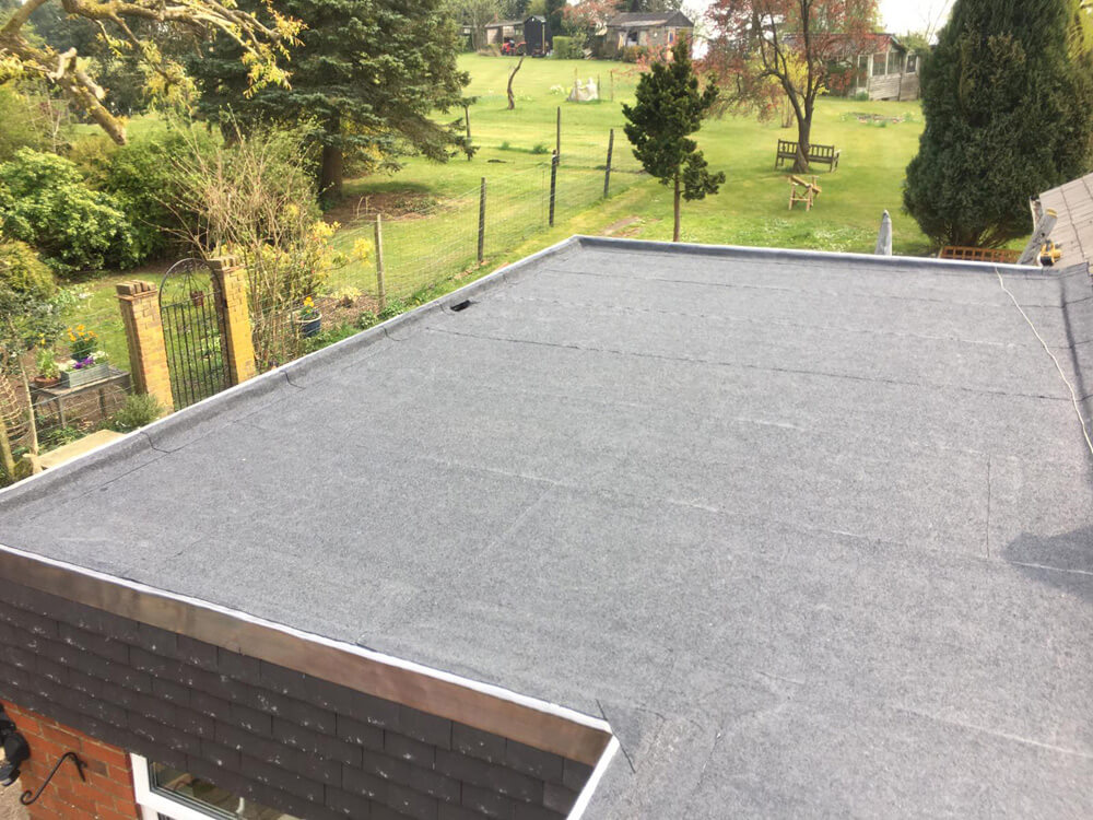 A high-angle view of a professionally installed grey felt flat roof with lead flashing, overlooking a large green garden with trees and a distant shed.