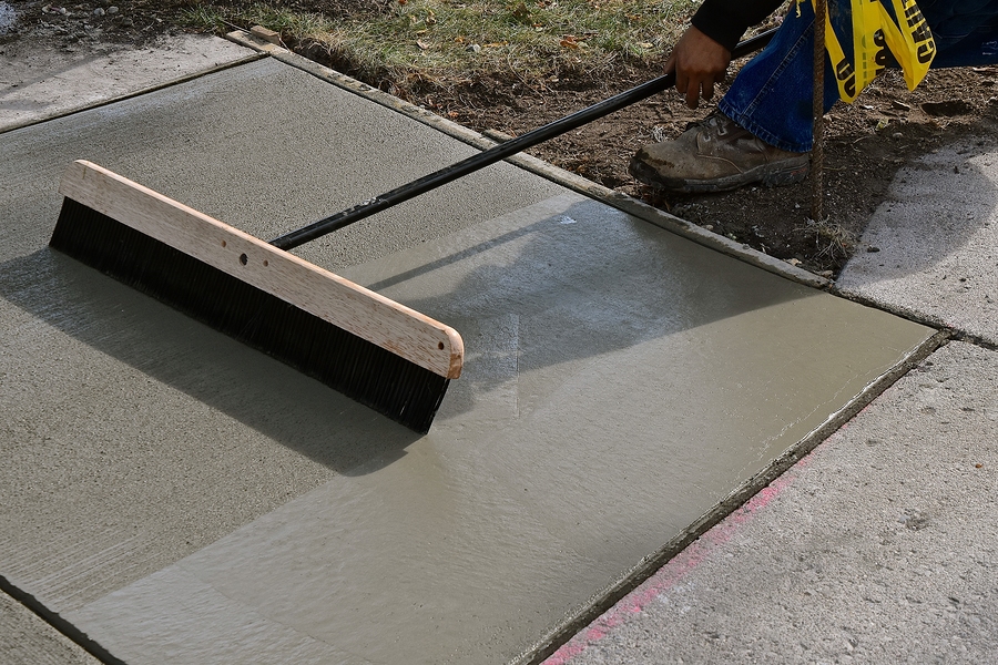 A concrete contractor uses a large broom to texture freshly poured concrete on a sidewalk.